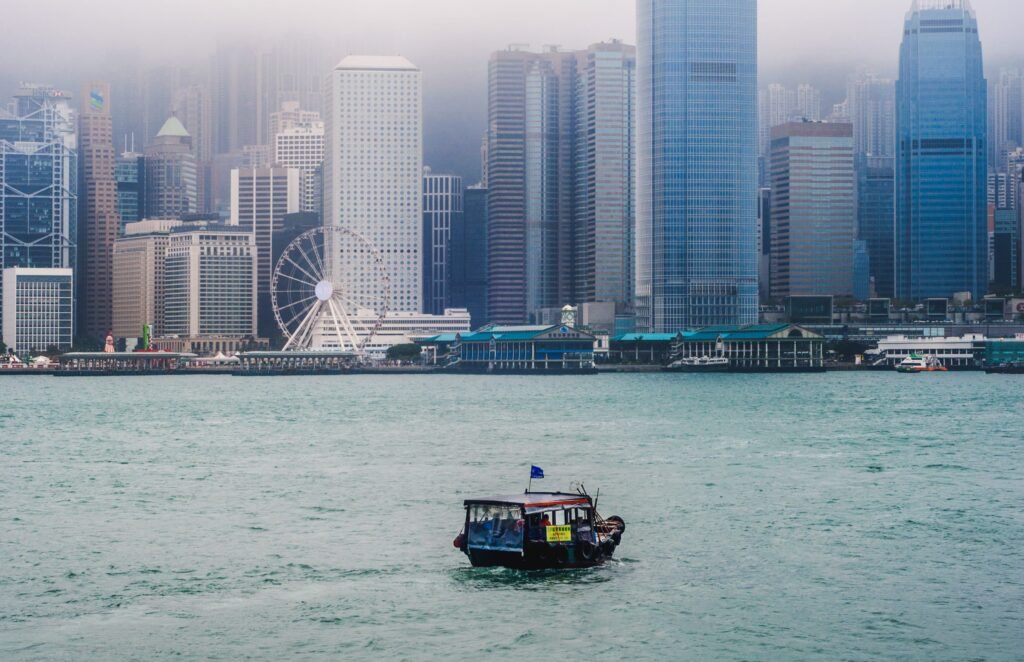 A small, traditional sampan boat floats alone in the waters of Victoria Harbour, dwarfed by the towering skyscrapers of Hong Kong Island in the background. The city skyline appears hazy, suggesting a sense of distance or isolation.