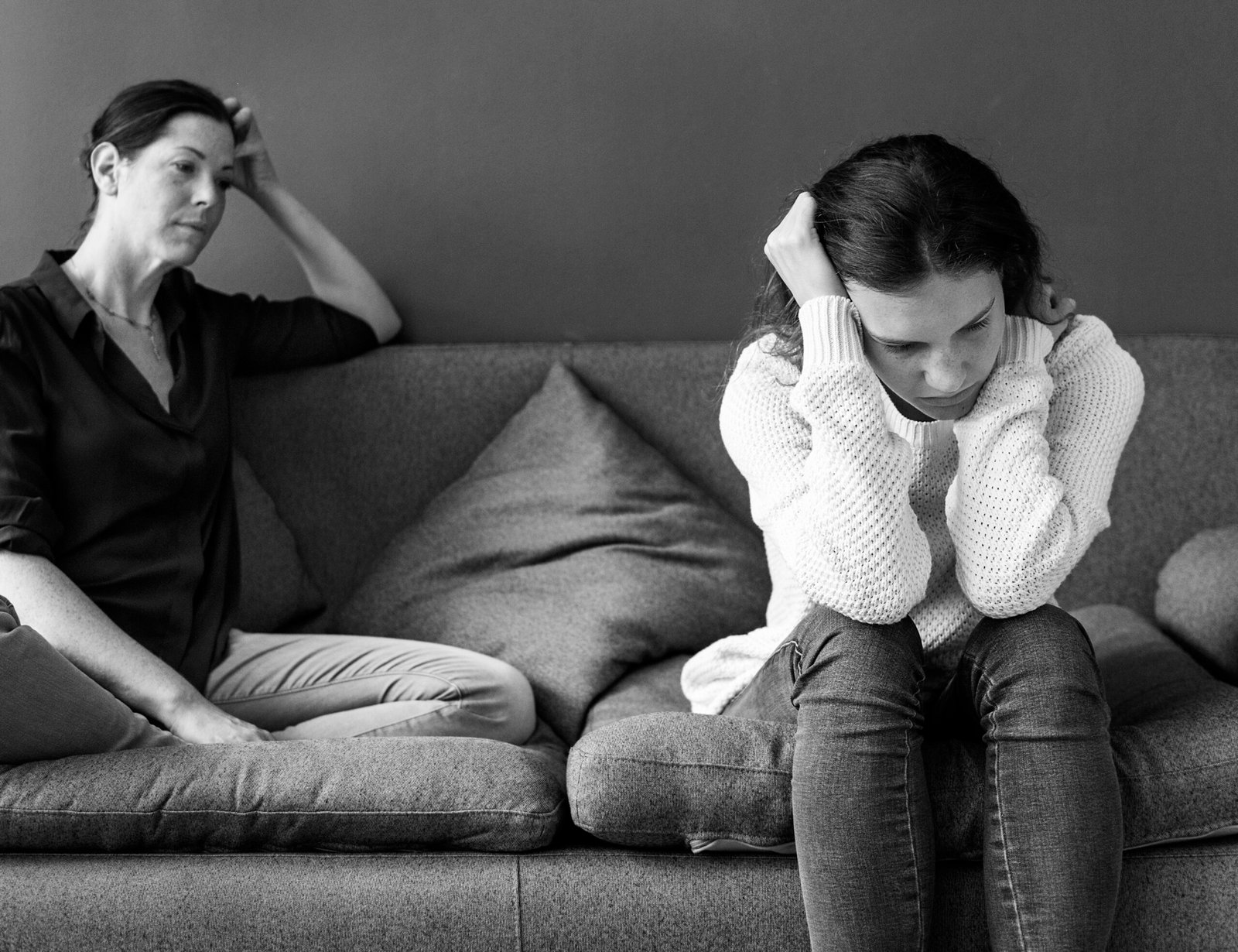 A mother and daughter sit on a couch, their bodies turned slightly away from each other. The daughter, in the foreground, hugs her knees and looks downwards, her expression obscured. The mother, in the background, leans back with one hand on her head, appearing concerned or distressed. The distance between them and their withdrawn postures suggest emotional distance or strain within their relationship. This image represents the relational aspect of eating disorders, highlighting how family dynamics and difficulties in relationships can contribute to these struggles.