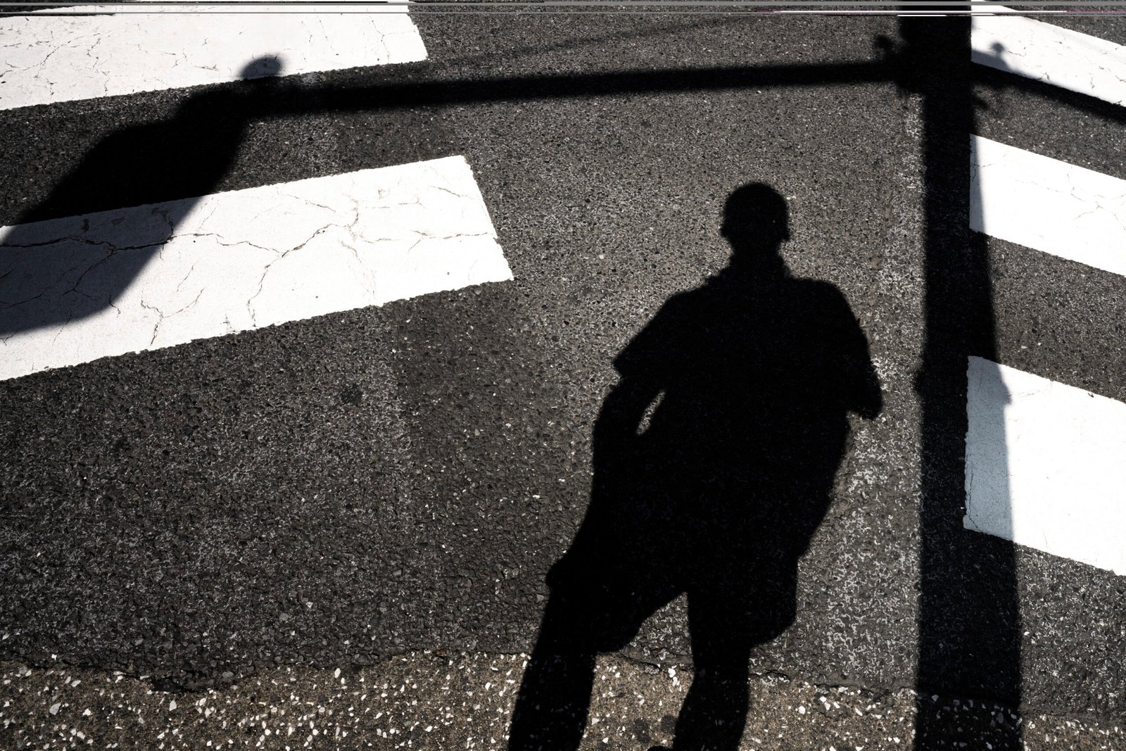 A distorted shadow cast on a crosswalk symbolizes the disconnect between self-perception and public image, a central characteristic of imposter syndrome.