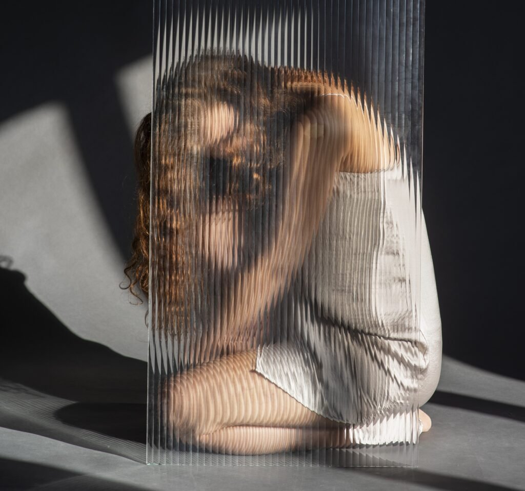 A person is crouched behind a textured glass panel. The glass distorts their figure, creating a fragmented and obscured image. The person's head is bowed, and their body is curled inwards, conveying a sense of vulnerability and emotional distress. The lighting casts shadows, adding to the feeling of isolation and hidden struggle. This image represents the internal struggles and emotional distress that can manifest as physical symptoms in psychosomatic issues, highlighting the complex mind-body connection.