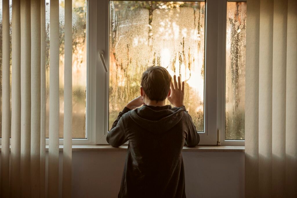 Person standing indoors with hands on a foggy window, looking outside, symbolizing emotional isolation and the desire for wholeness and connection.