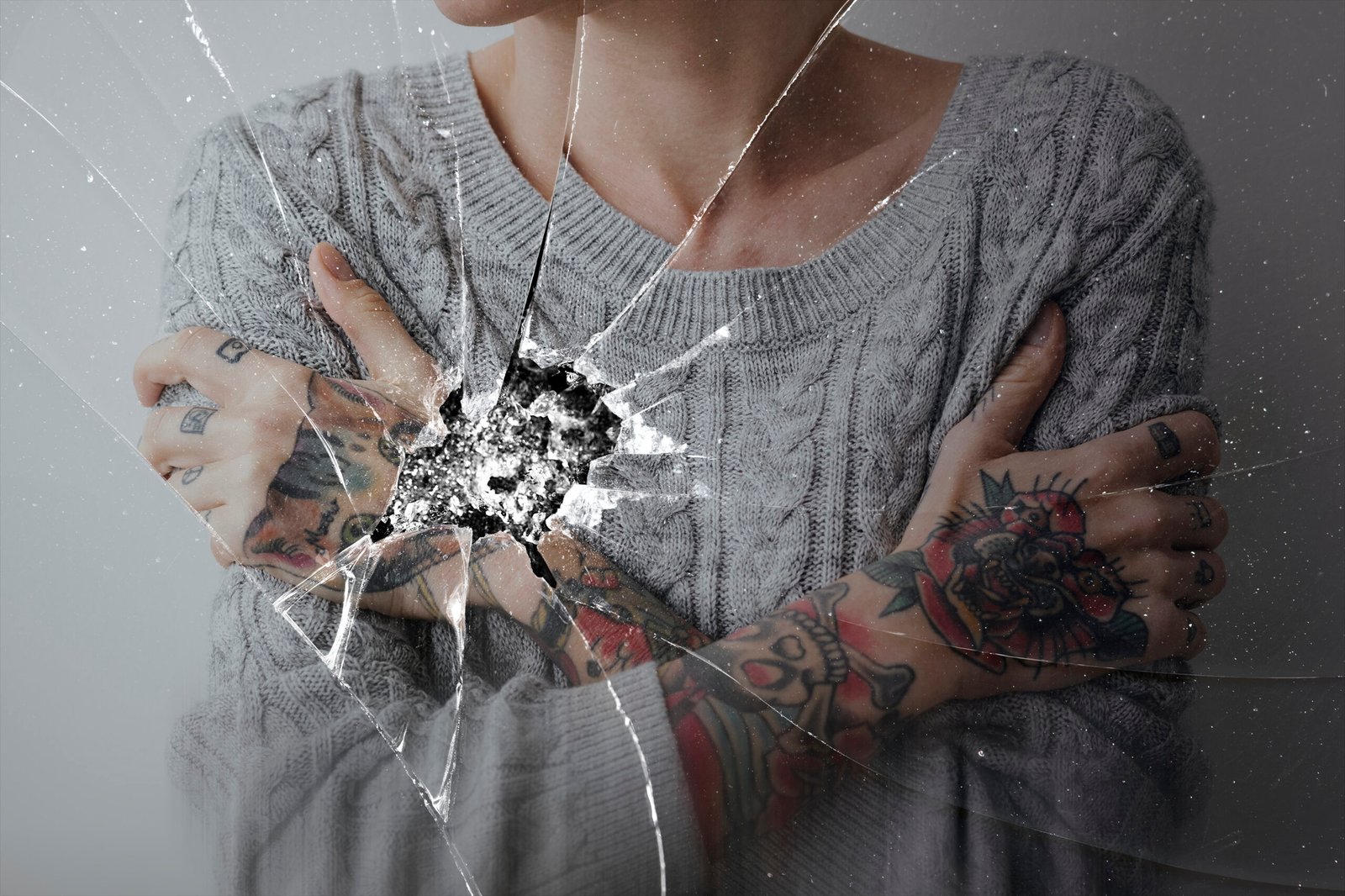 A person wearing a knitted sweater with arms crossed over their chest, holding themselves protectively. The image is overlaid with the effect of a cracked mirror, with the largest crack centered on their chest, symbolizing emotional vulnerability and inner conflict. mirror stage