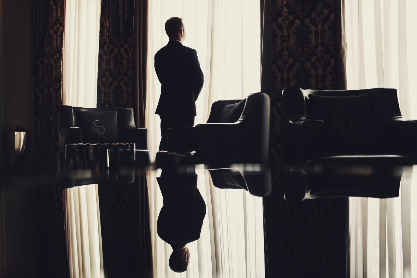 Silhouette of a man in a dark suit standing with his back to the camera in front of a bright window, reflected on a polished floor in a luxurious, somber room. A visual representation of a 'sealed life' and the fear of intimacy.